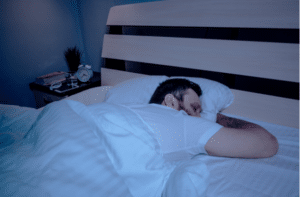 A lonely bearded dark-haired man sleeps soundly lying on his stomach at night on the bed, hands clasped.