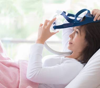 Woman in white clothes laying with knees up in bed hands holding and wearing CPAP mask