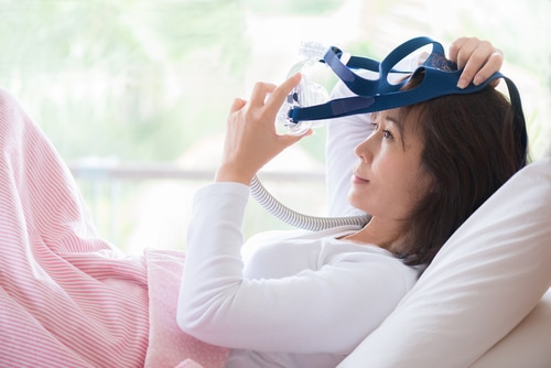 Woman in white clothes laying with knees up in bed hands holding and wearing CPAP mask