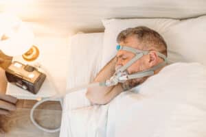 Man looks unwell and unhappy in bed trying to sleep with a CPAP machine for sleep apnea