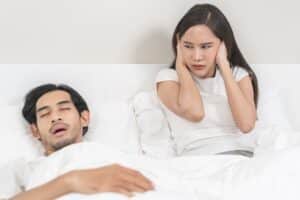 Couple in bed with the woman covering her ears, visibly annoyed by her husband's loud snoring, illustrating sleep apnea, sleep problems, and the impact on relationship health