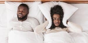 Frustrated woman covering her ears with a pillow while partner snores loudly beside her in bed